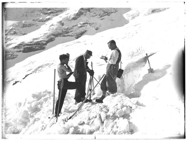 Scan einer Fotografie von Wilhelm Paulcke (600x455px, SW): Drei Männer auf einem schneebedeckten Hang (Gletscher). Der Mann links hält eine Kamera, der Mann in der Mitte ein Stativ, der Mann rechts einen Notizblock mit Stift.