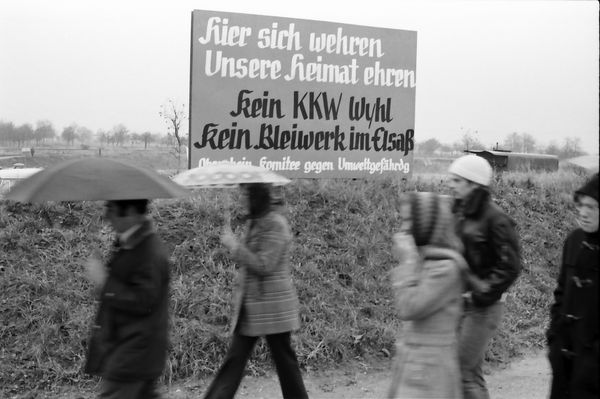 Schwarz-weiß-Fotografie einer Kundgebung gegen den Bau eines Atomkraftwerks im badischen Wyhl, 10. November 1974. Oben mittig ein Schild mit der Aufschrift: Hier sich wehren. Unsere Heimat ehren. Kein KKW Wyhl. Kein Bleiwerk im Elsaß. [Oberrheinisches] Komitee gegen Umweltgefährd[un]g. Im Vordergrund unten fünf vorbeilaufende Personen, zwei davon mit Regenschirm.